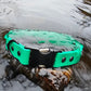 A green dog collar with an Alu-max buckle, placed on a rock surrounded by flowing water.